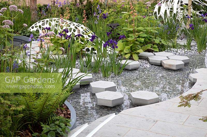 View of sculptures and cottage garden planting, with hexagonal stepping stones over the pond, bordered by centranthus ruber 'Albus', rodgersia, and iris sibirica. Garden: The Manchester Garden. Chelsea Flower Show 2019.
