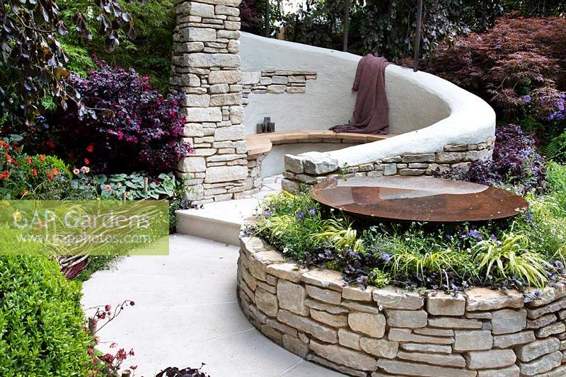 Curved seating area in Purbeck stone, enclosed in purple beeches, acers and herbaceous planting. Garden: The Kingston Maurward Garden. Sponsors: Miles Brown, Kingston Maurward College, Goulds Garden Centre, Wilks Landscaping, Holme for Gardens, The Green Gardener. Chelsea Flower Show 2019.