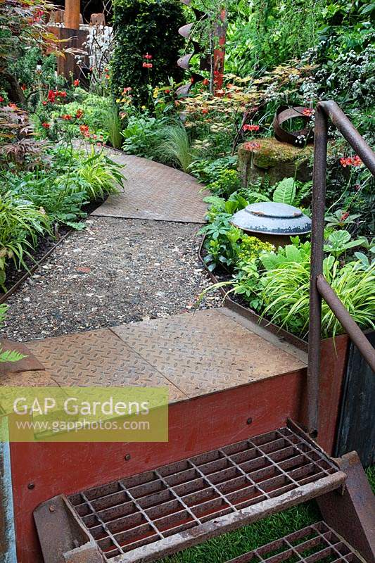 View of path made of rusty metal and porous spoil from the quarry, with metal steps, bordered by geum 'Totally Tangerine' and mixed grasses. Garden: Walker's Forgotten Quarry. Sponsor: Walkers Nurseries. Chelsea flower show 2019.