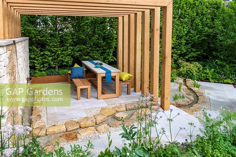 Kampo No Niwa Garden. A wooden pergola and stone patio provide a place to view the garden filled with medicinal plants. A rill winds its way past, inspired by the Hokkaido region of Japan. Sponsor: Kampo no Niwa. RHS Chelsea Flower Show 2019.