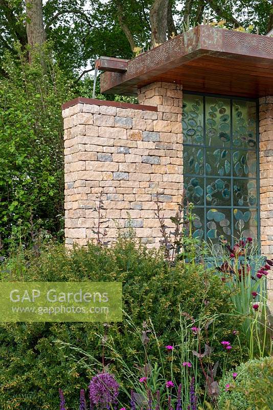 Natural stone building with green roof and water feature and hand-crafted glass panels by Wendy Newhofer, Taxus baccata dome in foreground - The Warner's Distillery Garden, RHS Chelsea Flower Show 2019.