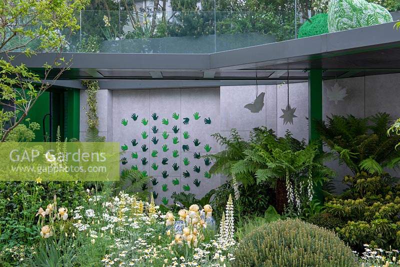 Balcony providing some shade to ferns and white foxgloves, plants in foreground include Iris, Lupinus 'Polar Princess', Orlaya grandiflora and Pittosporum tobira -  The Greenfingers Charity Garden, RHS Chelsea Flower Show, 2019.