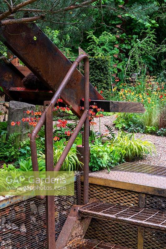 Industrial style rusted steps leading up to a gravel path, through orange and green planting - The Walker's Forgotten Quarry Garden, RHS Chelsea Flower Show 2019