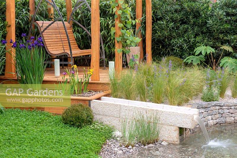 Swinging bench next to a water feature, Clover lawn and bed full of Stipa tenuissima and Nepeta - The Harmonious Garden of Life, RHS Chelsea Flower Show 2019.