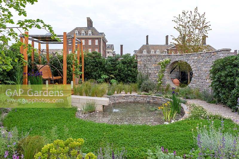 Clover lawn next to a circular pool with dry stone wall, platform with swinging bench and bed of Stipa tenuissima and Nepeta - The Harmonious Garden of Life, RHS Chelsea Flower Show 2019.