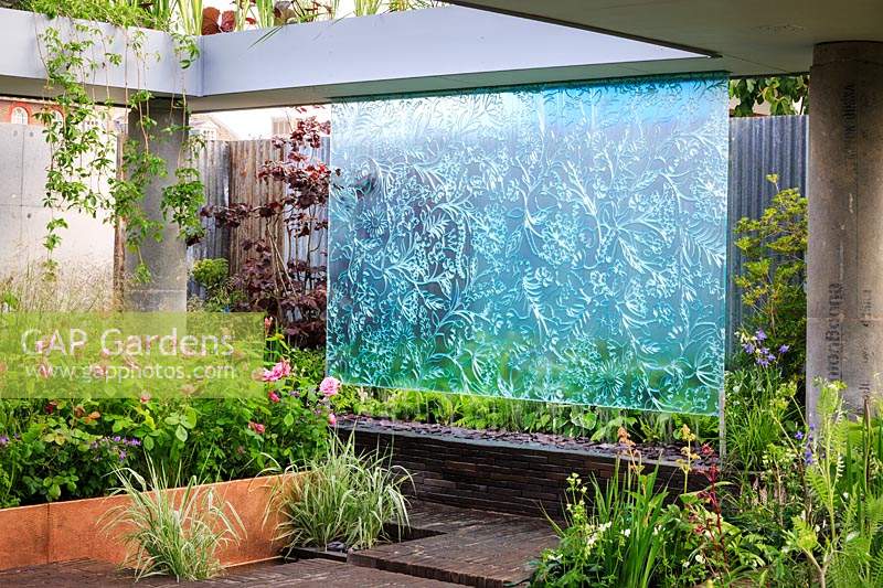 The Silent Pool Gin Garden. Etched water wall surrounded by a concrete structure imitating an urban landscape. Two plant modules within the garden generate electricity from the living plants. Plants in the hammered copper raised bed Rosa 'Gertrude Jekyll' and Deschampsia cespitosa. Behind the glass screen is a Corylus maxima 'Purpurea' Purple Hazel. Phalaris arundinacea var. picta 'Feesey' sits in the water of the geomtric pond and on the margins. Other plants include Rodgersia podophylla and Geranium. Sponsor: Silent Pool Distillers