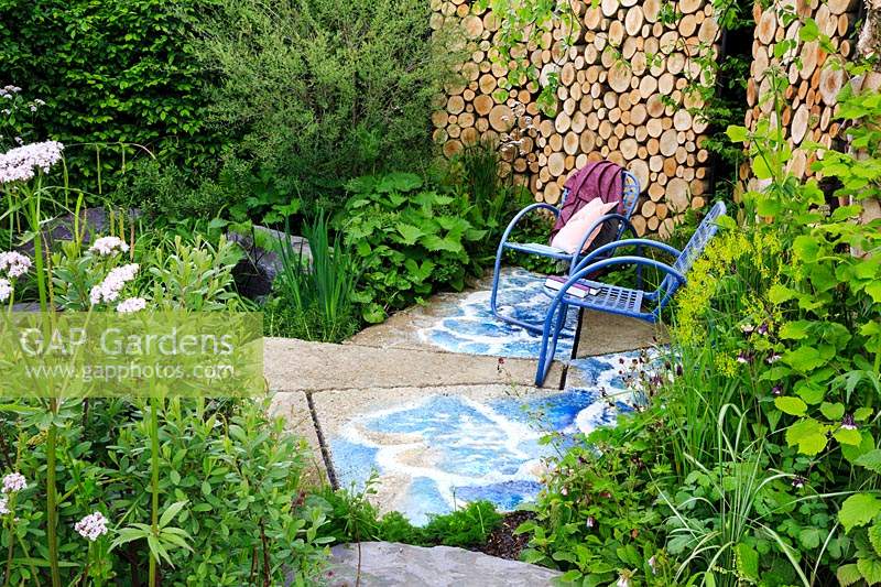 Viking Cruises: The Art of Viking Garden. Sunken sitting area featuring graffitied concrete slabs and blue metal chairs. Cut log-end wall behind. Plants include: Valeriana officinalis, Myrica gale, Geum rivale, Symphytum x uplandicum, Aquilegia, Carex - Sedge, Corylus avellane - Hazel and Salix - Shrubby Willow. Sponsor: Viking Cruises. 