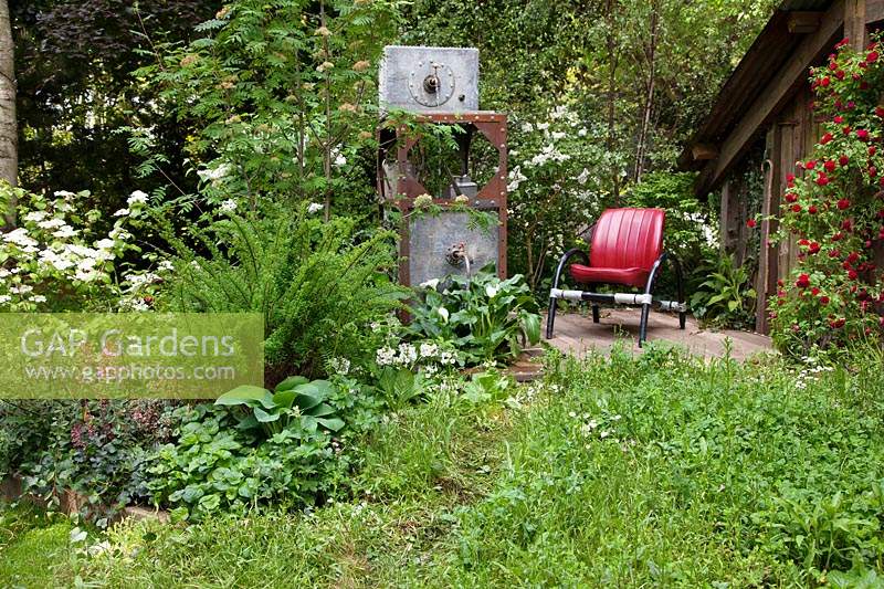 The High Maintenance Garden for Motor Neurone Disease Association, view of garden where old metal containers have been recycled into a water feature and where the planting includes red rambling roses, Hosta, Viburnum and Primula - Sponsor: MND Association.