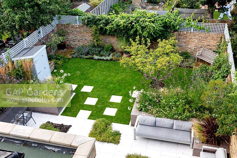 View of garden from second floor of house. Looking out on lawn with white stone paving and patio with seating area. Raised beds full of mixed planting, garden enclosed by brick walls topped with trellis.