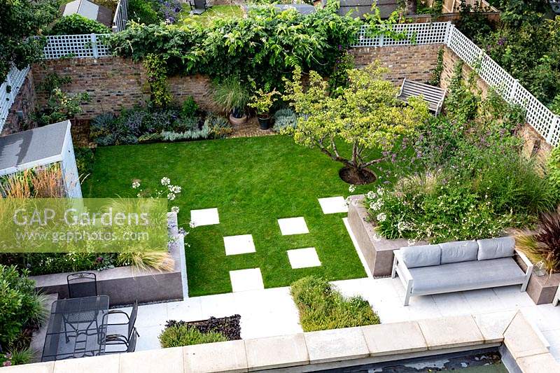 Overview of garden from second floor, looking over lawn with white stone paving and patio, with seating areas. Raised beds filled with mixed plantings. Garden surrounded by brick walls topped with trellis.
