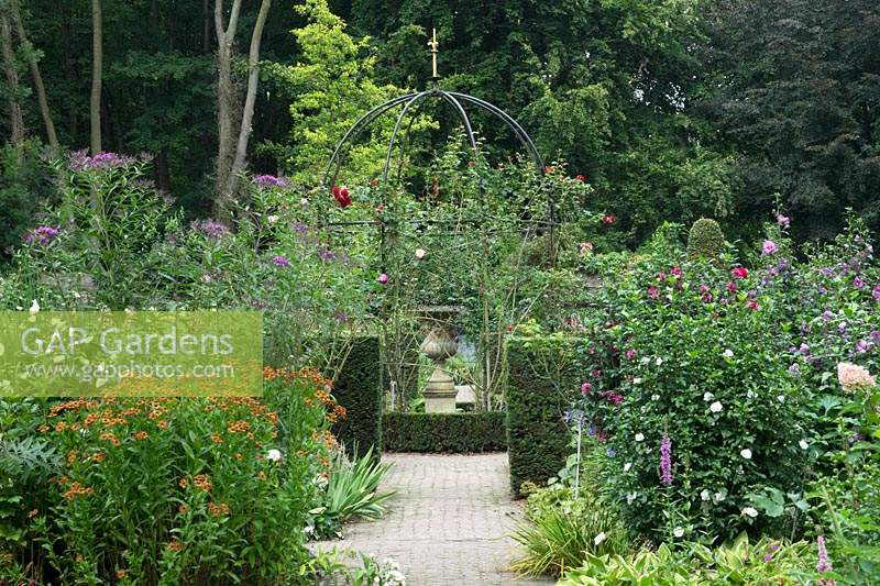 Flower beds either side of path leading to metal arbour with Rosa - Climbing Rose - and urn on pedestal 
