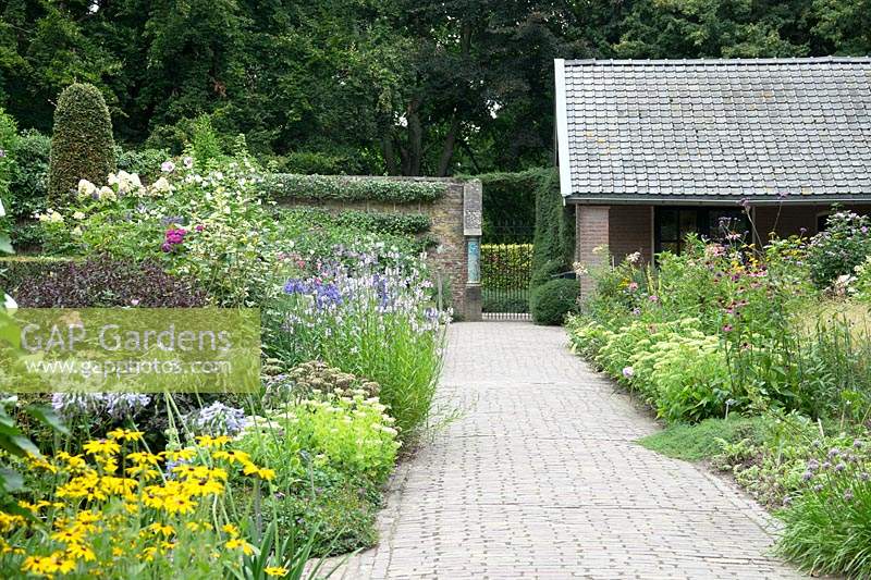 View along paved path with flowerbeds either side towards entrance. Flower beds contain: Hydrangea, Allium, Hibiscus, Rudbeckia and Echinacea