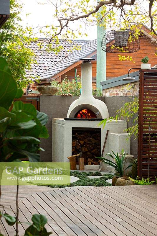 A bespoke cement rendered pizza oven in the back corner of a backyard showing a slat hardwood timber screen and a timber deck.