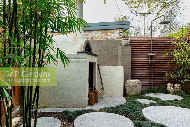 Pizza oven in back corner of garden, next to a painted brick fence, a timber slat screen with an outdoor shower, a stone carved Indonesia pot, a family group of sculptures, a large glazed water bowl, semi rustic stone pots, concrete circular cast in situ stepping stones in amongst a thick planting of mini Mondo grass.