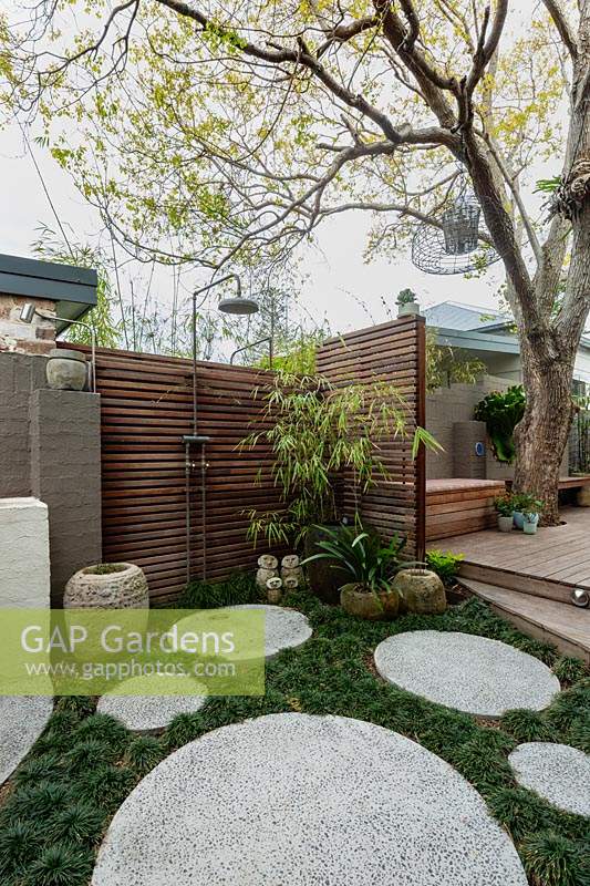 Corner of a garden next to a painted brick fence, featuring a timber slat screen with an outdoor shower, a stone carved Indonesia pot, a family group of sculptures, a large glazed water bowl, semi rustic stone pots, concrete circular cast in situ stepping stones in amongst a thick planting of mini Mondo grass.