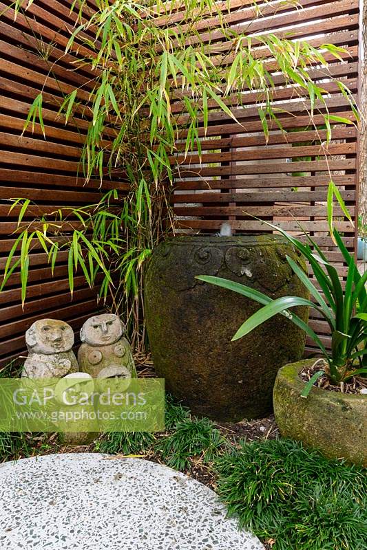 Detail of a family group of hand carved sculptures in front of a timber slat screen with a carved stone pot water bowl, set in amongst a planting of mini Mondo grass next to an in situ cast concrete stepping stone.