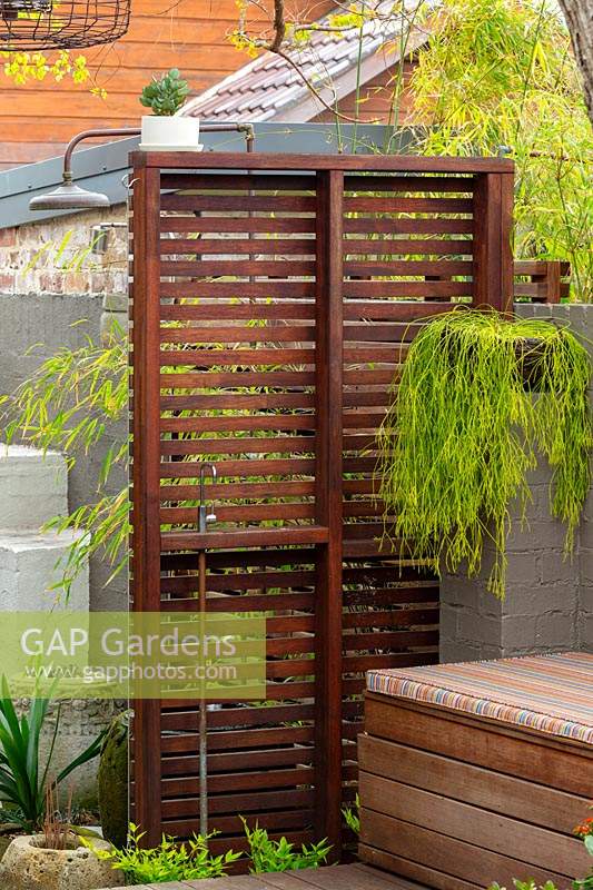 Freestanding hardwood timber slat screen with an inbuilt cold water tap next to a timber bench seat infront of a painted brick fence, featuring a Rhipsalis, succulent in a hanging basket.