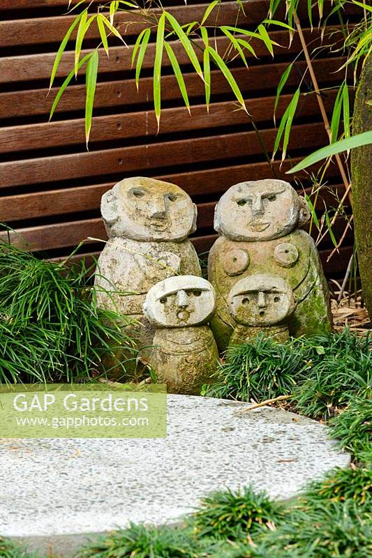 Detail of a family group of hand carved sculptures in front of a timber slat screen set in amongst a planting of mini Mondo grass next to an in situ cast concrete stepping stone.