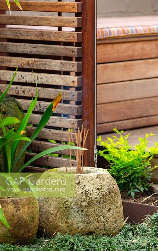 Semi rustic stone pot used as an incense burner next to a pot of Clivea and a thick planting of mini Mondo grass in front of a timber slat screen.