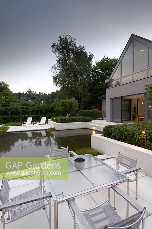 View across dining area to recliners on paved patio surrounded by pool in modern garden. 