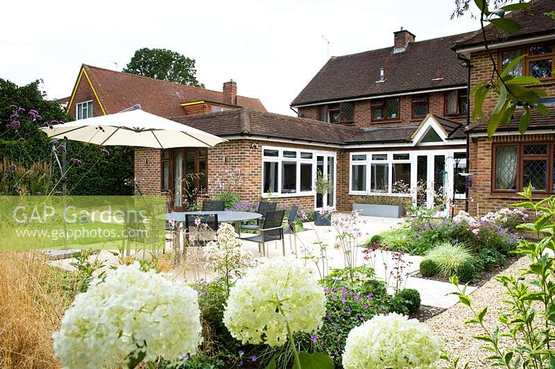 View across patio with small beds of perennials and dining area, towards back of the house with patio doors and brick extension