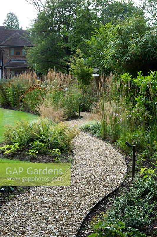View along curving gravel path with retaining edge towards house. Perennial beds on either side with ornamental grasses 
