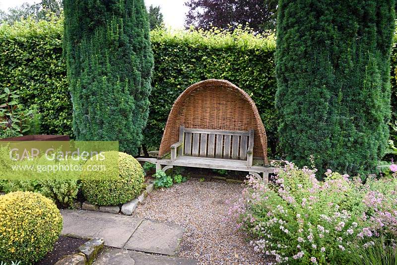 Clipped golden box, fastigiate yew and herbs including lemon balm and thyme frame a bench with woven willow hood in the garden at York Gate, Adel in July.