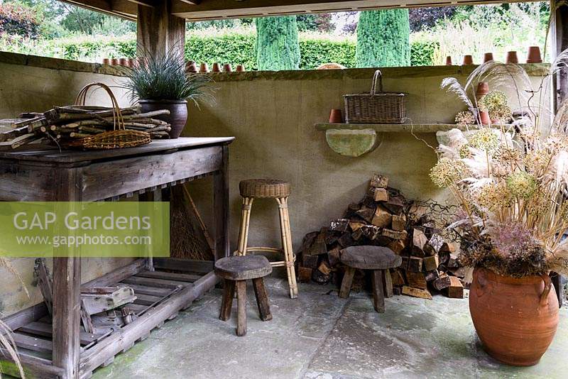 Covered open-sided potting shed with wooden stools, basket work, terracotta pots and arrangement of dried seedheads at York Gate Garden, Adel in July.