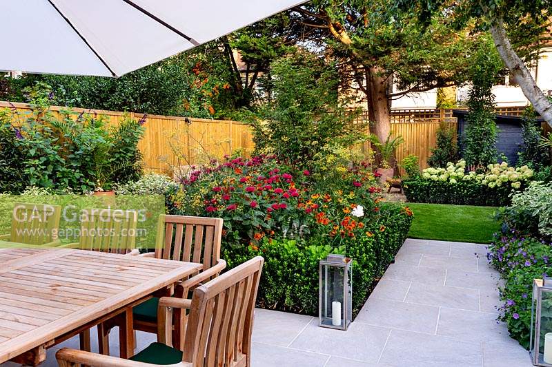 Stone patio area in West London garden, with wooden table, chairs and parasol. In the background there is a border featuring Monarda Fire Ball, Acer palmatum, Helenium Moerheim Beauty, Euonymus Jean Hughes.