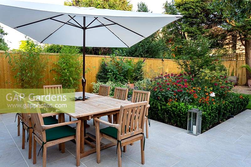 Stone patio area in West London garden, with wooden table, chairs and parasol. In the background there is a border featuring Monarda Fire Ball, Acer palmatum, Helenium Moerheim Beauty, bamboo in terracotta containers, Euonymus Jean Hughes.