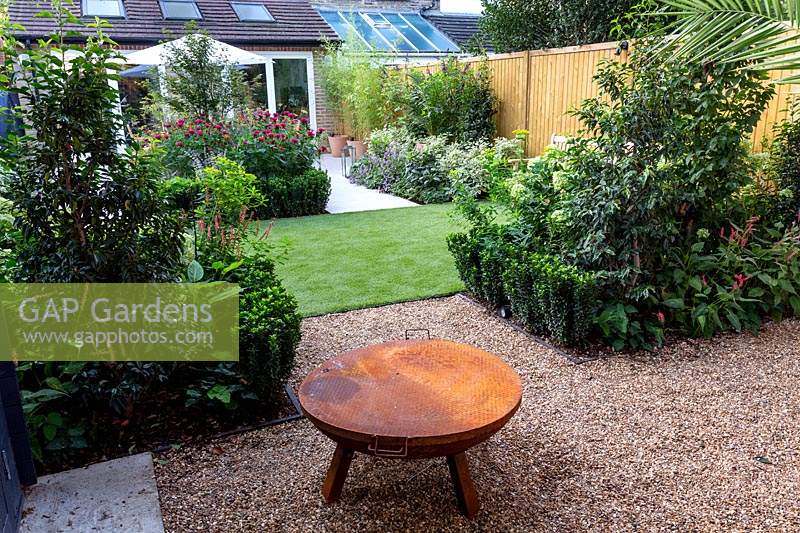 Gravel patio with fire pit in West London garden, with artificial lawn - View towards house - planting includes Persicaria Orange Field, Euonymus Jean Hughes, Monarda Fire Ball, Cornus alba Sibirica Variegata.