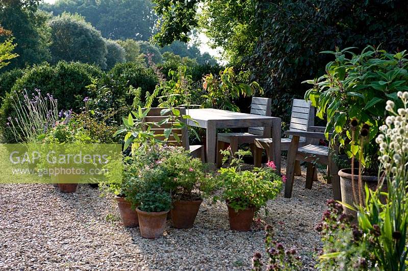 Potted plants, wooden table and chairs in gravel area of garden. 