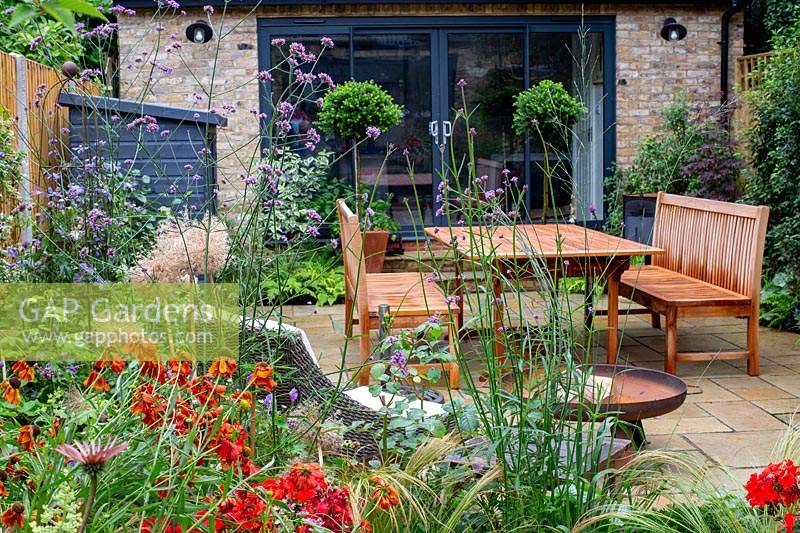 Contemporary garden in West London with stone patio and herbaceous borders, with view towards wood table, chairs and garden room. Planting includes Verbena bonariensis, Helenium Moerheim Beauty, Nassella tenuissima,