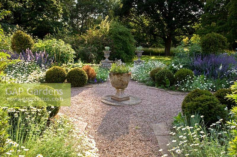 Central stone container in formal gravel garden, surrounded by symmetrical borders of flowering perennials and clipped Buxus - Box - balls