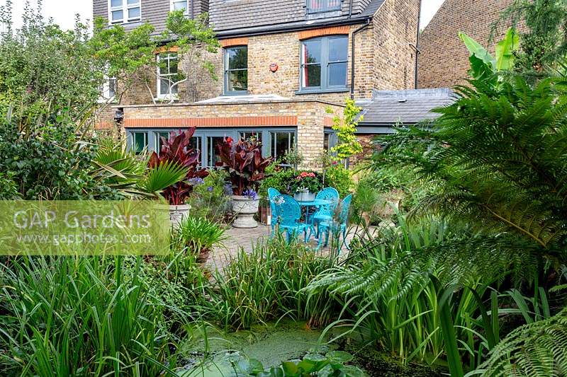 Modern cottage garden in West London. Looking towards house including pond area and patio with blue seating and table.