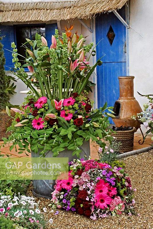 The Climate Change Garden - mixed flower arrangments with Gerbera on gravel in front of building with thatch roof and blue door