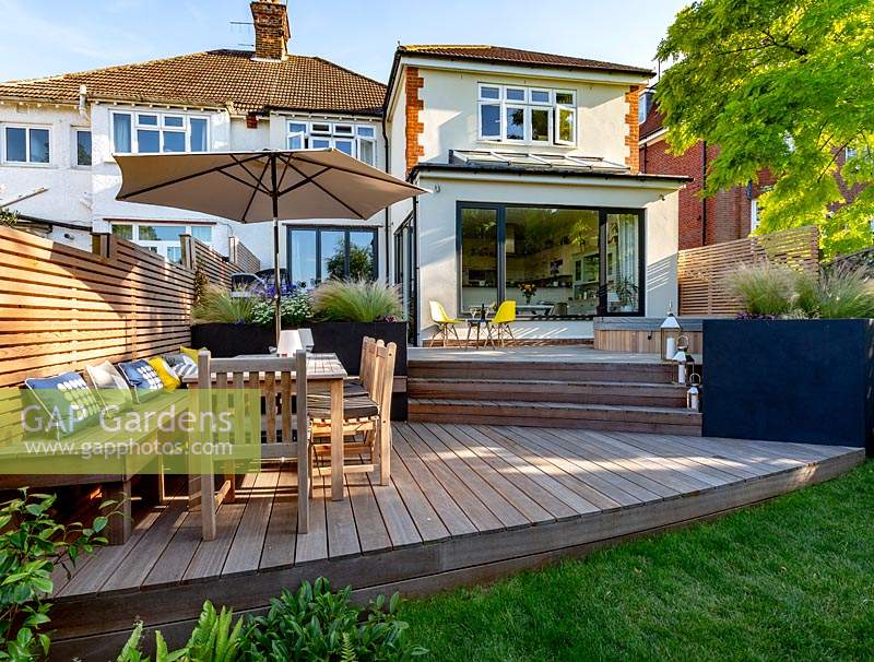 Contemporary garden in Wimbledon with decking on two levels. The lower section includes a bespoke built in bench with cushions, a table with parasol and chairs. In the background are grey raised beds with perennials.