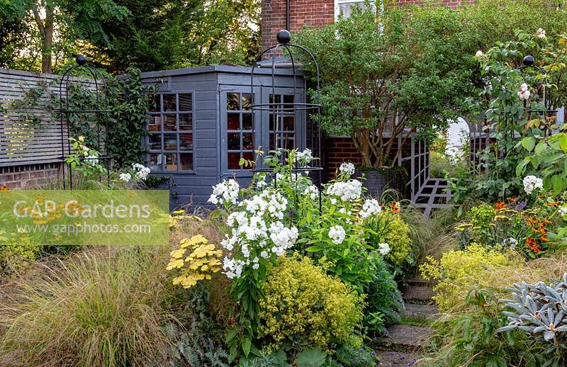 Small garden, planting includes: Phlox 'David', Achillea 'Moonshine', Stipa arundinacea, Helenium 'Moerheim Beauty' and Alchemilla mollis. House wall disguised by summerhouse, tree in large pot and mirror surrounded by trellis to create trompe l'oeil