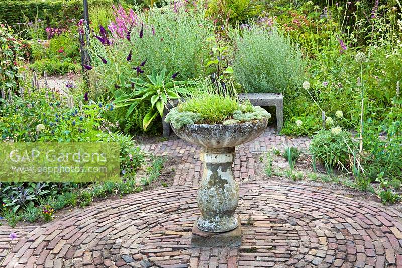 Herb beds around a circular brick paved area with central stone bird bath planted up 