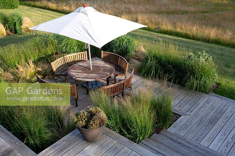 Meadow garden in Aldeburgh, Suffolk. High view of decked patio area with wooden table with parasol and chairs. Herbaceous border planting includes: Panicum virgatum - Heavy Metal, Molinia caerulea subsp. arundinacea - Transparent, Stipa tenuissima, Hakonechloa macra.
