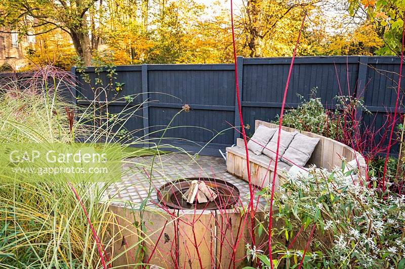 Seating area with fire pit and round wooden bench surrounded by Miscanthus sinensis 'Morning Light' - eulalia and Cornus alba 'Sibirica'