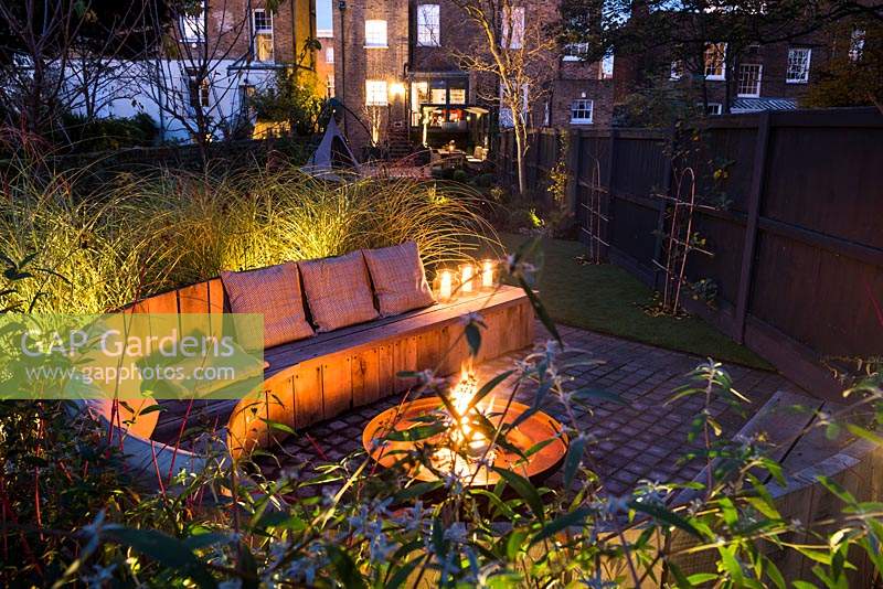 Seating area with fire pit and round wooden bench surrounded by Miscanthus sinensis 'Morning Light' - eulalia and Cornus alba 'Sibirica' Siberian dogwood AGM at night 