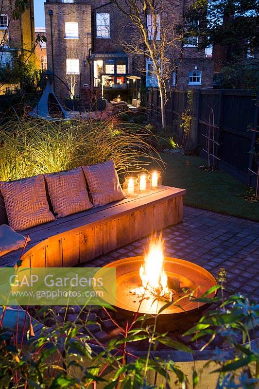 Seating area with fire pit and round wooden bench surrounded by Miscanthus sinensis 'Morning Light' -Eulalia and Cornus alba 'Sibirica' - Siberian dogwood, AGM at night in Autumn.