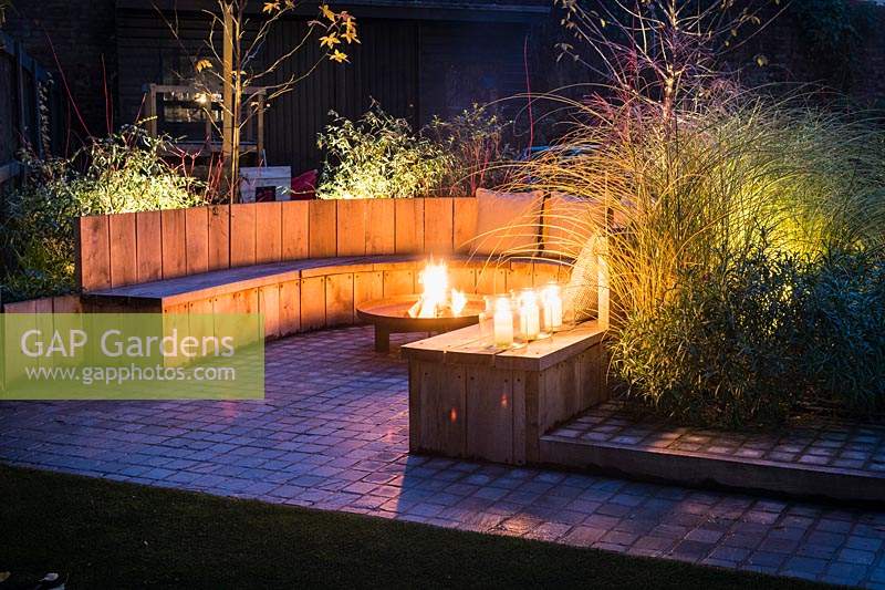 Seating area with fire pit and round wooden bench surrounded by Miscanthus sinensis 'Morning Light' - eulalia and Cornus alba 'Sibirica' - Siberian dogwood, AGM at night in Autumn.