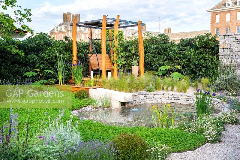 A garden with swing by the pond with water feature surrounded by ground covering plants. The Harmonious Garden of Life. RHS Chelsea Flower Show 2019
