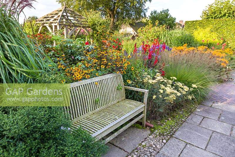 A wooden bench in The Lanhydrock Garden, noted for its hot colours. Planting includes: Rudbeckia, Achillea, Dahlias, Lobelia cardinalis, and Ligularia