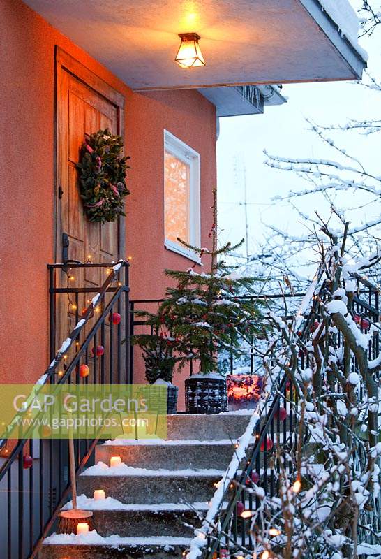 Winter decorations on steps by entrance door including lights, apples, wreath and christmas tree in pot