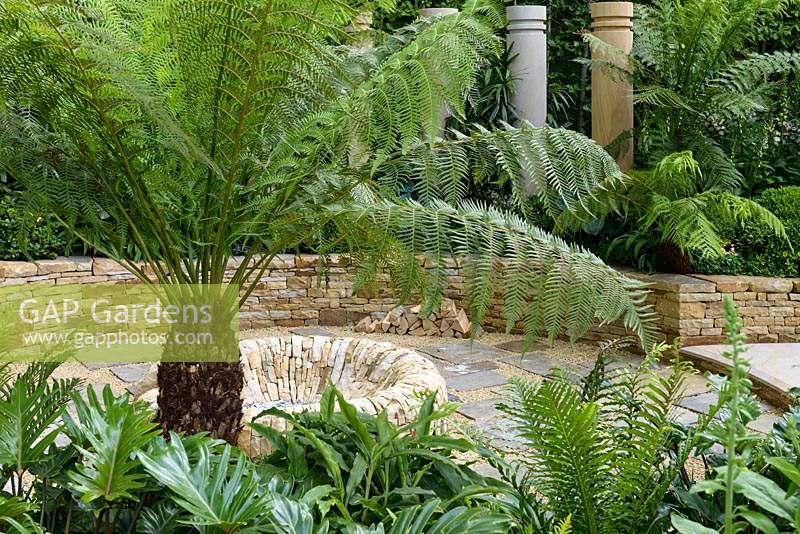 Dicksonia Antarctica Tree fern stands over circular stone sunken fire pit surrounded by stone columns - The Time In Between by Husqvarna and Gardena Garden at RHS Chelsea Flower Show 2015 - Sponsor: Husqvarna and Gardena