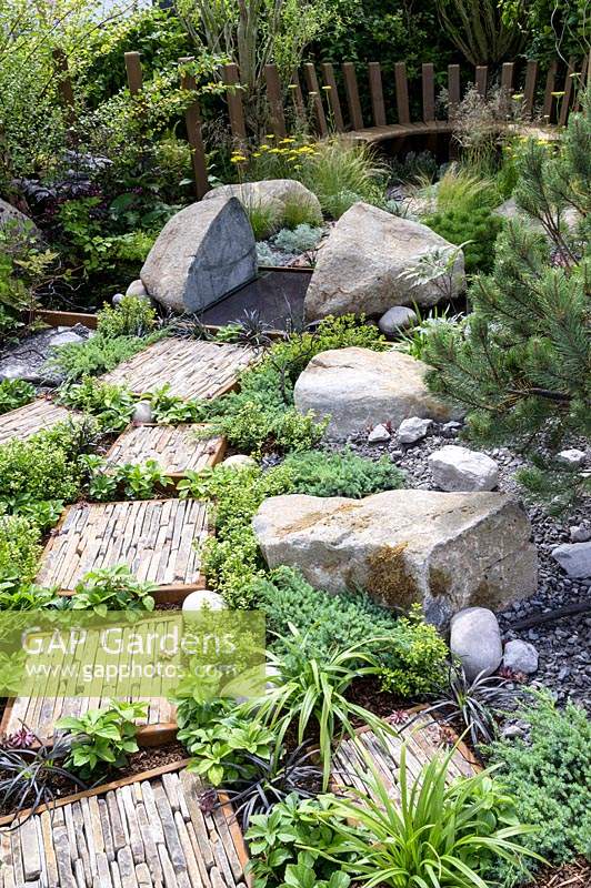 Natural rock and stone, with green foliage groundcover planting. Wooden seating and  stepping stones with slate pieces in corten steel frames with Deschampsia cespitosa, Achillea 'Moonshine', Ophiopogon planiscapus 'Nigrescens' - Through Your Eyes Garden - RHS Hampton Court Palace Garden Festival 2019. Sponsors: Kebony, CED Stone, R and G Metal Products, William's Art and Design, Practicality Brown.