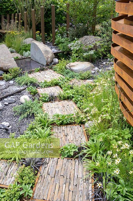 Natural rock and stone, with green foliage groundcover planting. Stepping stones with slate pieces in corten steel frames with Deschampsia cespitosa, Achillea 'Moonshine', Ophiopogon planiscapus 'Nigrescens' - Through Your Eyes Garden - RHS Hampton Court  Palace Garden Festival 2019. Sponsors: Kebony, CED Stone, R and G Metal Products, William's Art and Design, Practicality Brown.